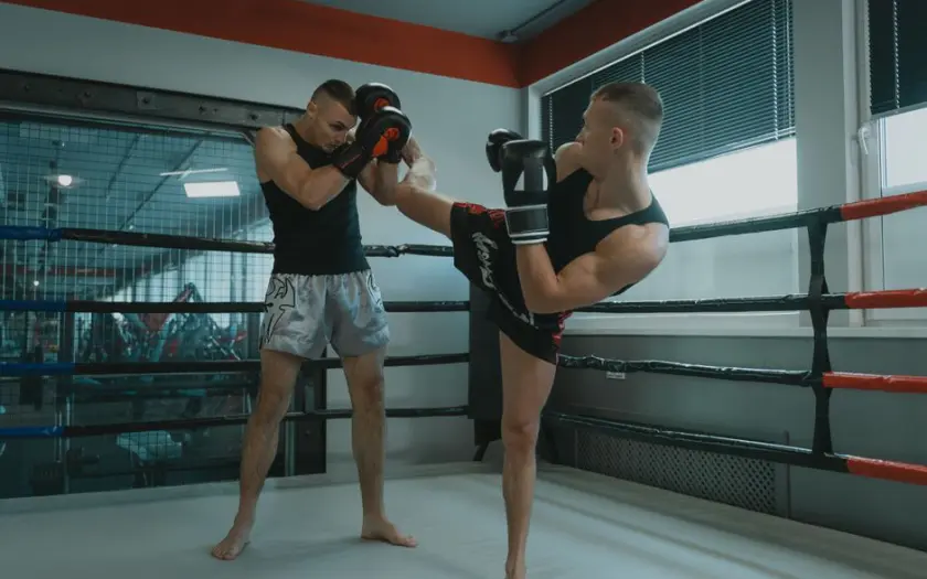 A powerful Muay Thai sparring bout between two muscular athletes in a gym boxing ring.