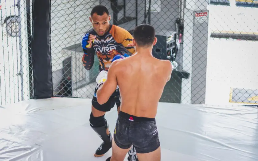 Two athletes engaged in a mixed martial arts sparring session inside a cage.