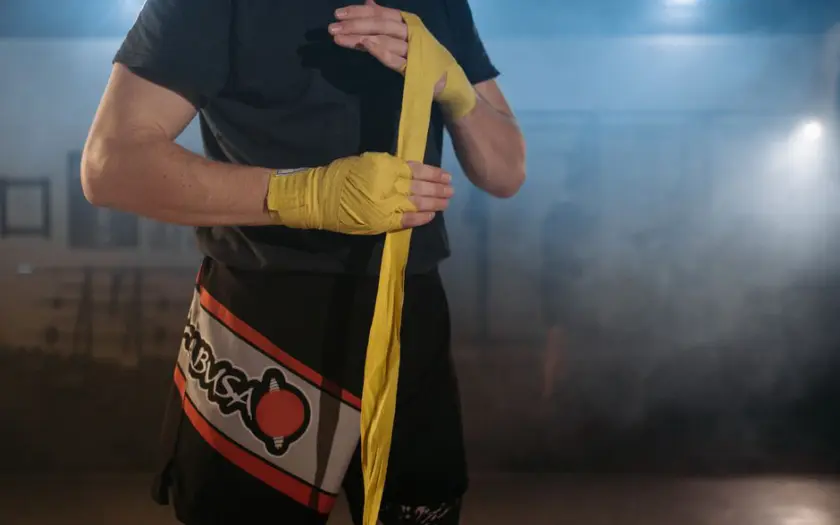 A close-up of an MMA fighter wrapping his hands with yellow bandages in a smoky gym setting.