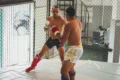 Two men engage in intense kickboxing sparring inside a gym arena.